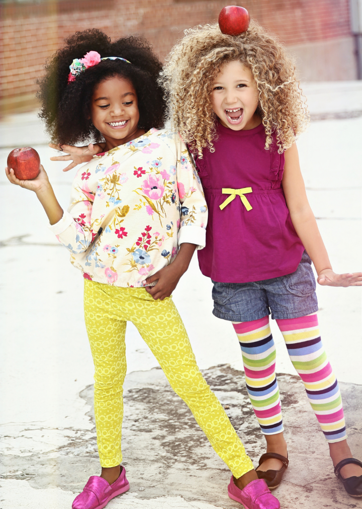 2 girls in bright color outfits standing together. One holds an apple, another has an apple on her head.
